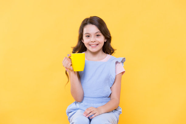 美味的奶茶。快乐的孩子捧着黄色背景的茶杯。小女孩喜欢喝茶。热饮。早上喝。早餐时间。好习惯。幸福是一杯茶图片下载