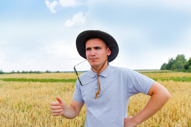 有趣的农民。收割机。农民站在金色的麦田里，背景是蓝天。一个戴着太阳镜和牛仔帽的年轻人在麦田里检查小麦。燕麦粮食生产图片下载