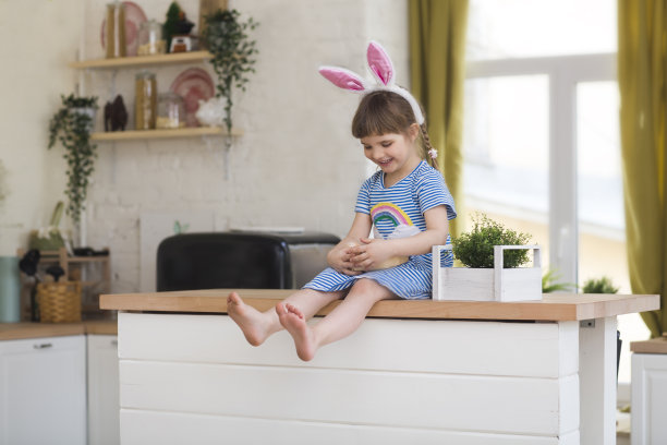 复活节快乐!快乐的家庭准备复活节。在房子的厨房里，一个戴着兔耳朵的可爱小女孩拿着一个画有鸡蛋的篮子。图片下载