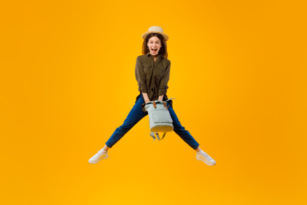 激动的女子抱着背包在半空中跳跃，黄色背景图片下载