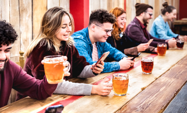 快乐上瘾的朋友在啤酒厂酒吧餐厅喝啤酒，玩手机的乐趣-生活方式的概念与千禧一代分享内容的技术设备-关注左女人图片下载