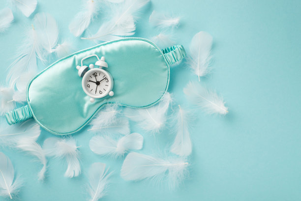 俯视图照片的蓝色丝绸眼罩与白色时钟上的许多白色嫩羽毛散落在柔和的天空蓝色孤立的背景空白空间图片下载