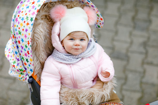 可爱的小女孩坐在婴儿车或婴儿车在寒冷的秋天，冬天或春天的一天。快乐微笑的孩子穿着暖和的衣服，时尚时尚的婴儿外套和帽子。雪落下来图片下载