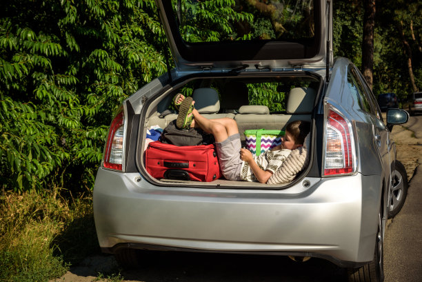可爱的小男孩躺在汽车后备箱的袋子和行李后面，准备去度假，带着快乐的表情。孩子们在玩智能手机图片下载