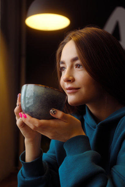 一个穿着蓝色连帽衫的漂亮女孩坐在咖啡桌旁。女人用一个大杯子喝咖啡。舒适的气氛。图片下载