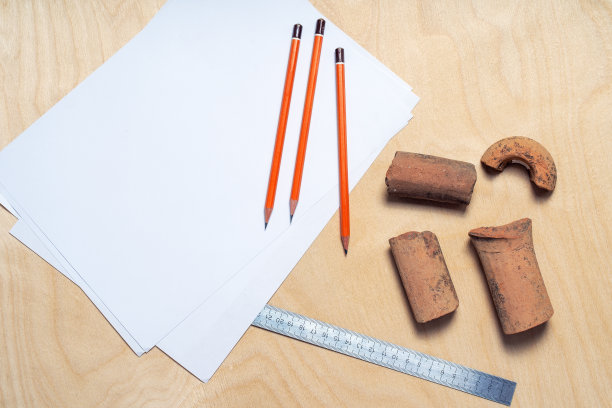 在考古发掘中发现的一些陶土文物，由于年代久远而受损，它们与纸张、金属尺子和铅笔放在木制背景上。图片下载