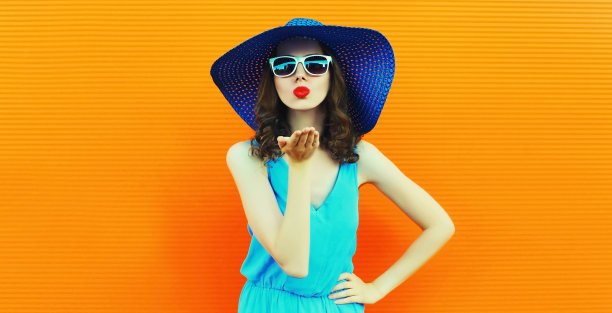 美丽的年轻女子的肖像吹她的嘴唇送甜蜜的空气吻戴在橙色背景的蓝色圆帽图片下载