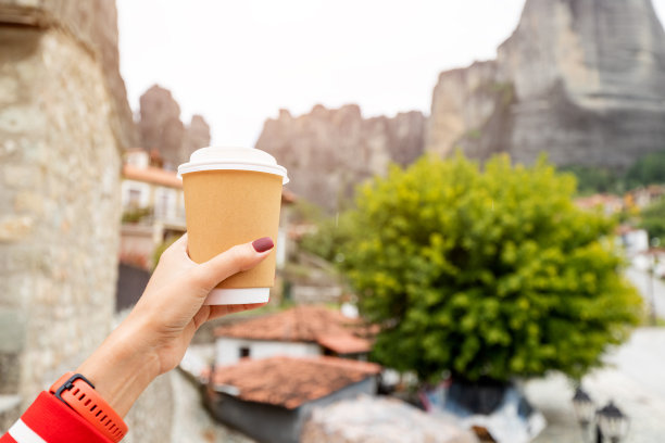 手拿一个带着新鲜热咖啡或茶的外卖纸杯，背景是著名的迈泰奥拉山脉图片下载