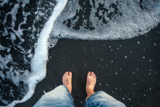 男人的脚在黑色的沙滩和白色的泡沫海浪。图片下载