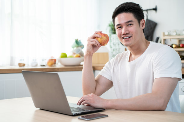 亚洲商人在家工作时吃健康沙拉的肖像。有魅力的帅哥用笔记本电脑上网学习，在厨房吃干净的蔬菜保健，看着相机。素材图片