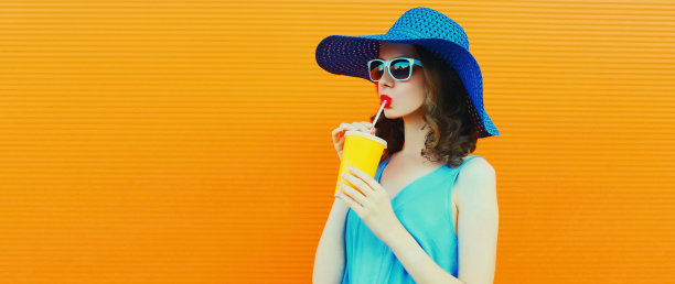美丽的年轻女子的肖像喝新鲜的果汁从杯戴夏季帽子在橙色的背景，空白的广告文字复制空间图片下载