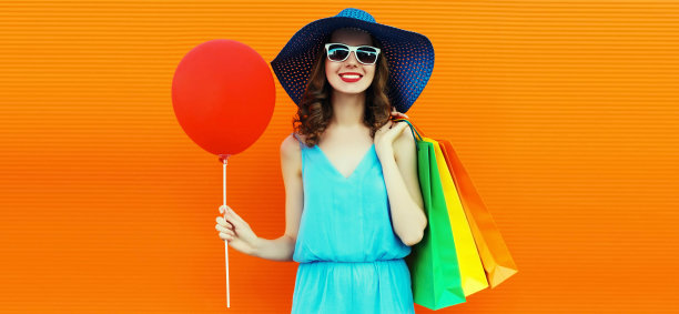 美丽的快乐微笑的年轻女子的肖像与彩色购物袋戴在橙色背景夏季草帽图片下载
