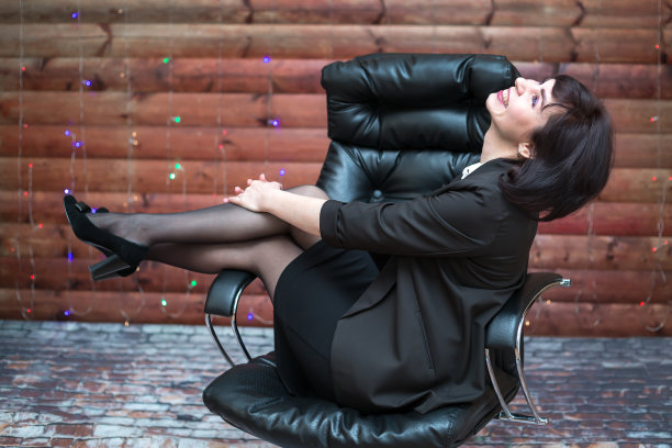 一位快乐的黑发女子坐在工作场所的皮椅上，双腿在木墙的背景下翘起图片下载