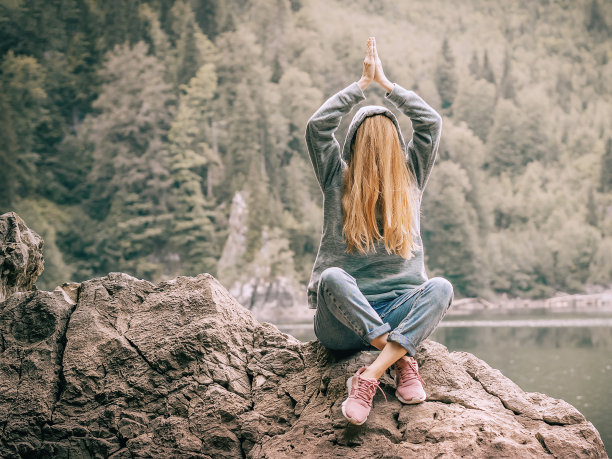 森林里的一个湖附近，一个女人坐在一块大石头上，双手交叉放在头上，把脸藏在头发下面。开玩笑的照片图片下载