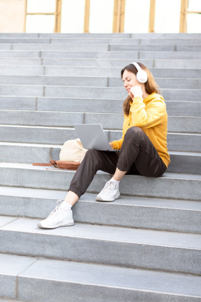 虚拟的日期。迷人的白种女人戴着无线耳机坐在楼梯的台阶上，用笔记本电脑在秋季户外视频通话，垂直图像，选定的焦点。图片下载