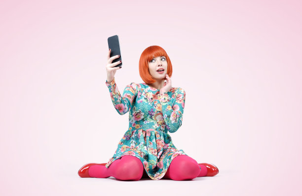 年轻漂亮的女孩穿着花裙子，手里拿着一个大的智能手机，背景是彩色的图片下载