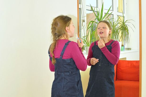 美丽时尚理念。孩子脸上有漂亮的笑容，在室内画嘴唇。小女孩拿着口红或睫毛膏站在房间的镜子前。孩子试着画嘴唇，用妈妈的化妆品化妆图片下载