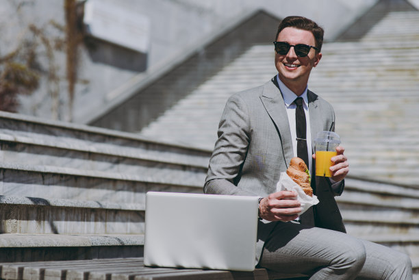年轻微笑的商人穿着灰色正装在笔记本电脑旁工作坐在市中心的水泥台阶上浏览互联网喝果汁吃牛角面包休息午餐在户外吃早餐。图片下载