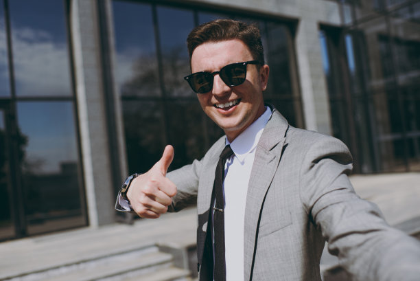 近距离拍摄年轻的员工，一名20多岁的商人，穿着灰色西装，走在市中心的办公室玻璃墙外，在社交网络上用手机自拍，展示胜利的姿态图片下载