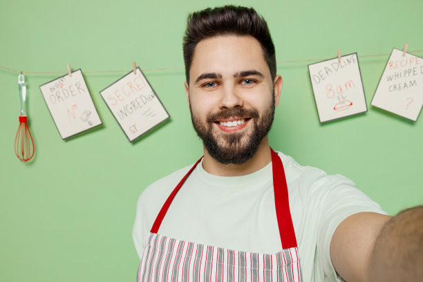 年轻微笑快乐的男厨师，糖果师，面包师，20多岁，穿着条纹围裙，正在用手机拍摄自拍，隔离在素色的淡绿色背景的工作室肖像。烹饪食物的概念。图片下载