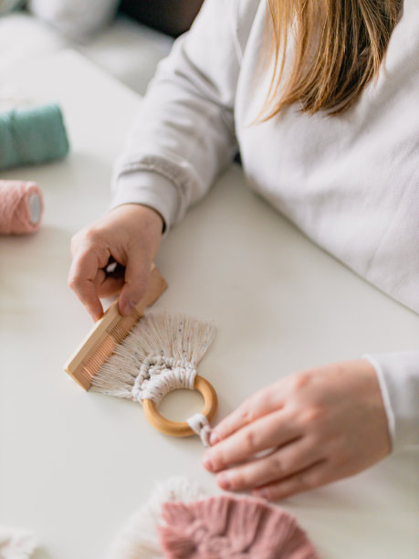 一名女子正在整理一件几乎完工的流线织物的最后细节，在一间家庭房间里梳理绳子。关闭了。天然棉线和木珠。女性的爱好。壁挂装饰。图片下载