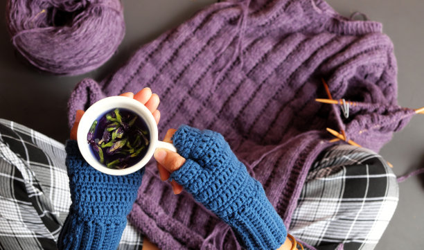 女人手戴蓝色连指手套钩针，在织紫色毛衣的同时端杯茶图片下载