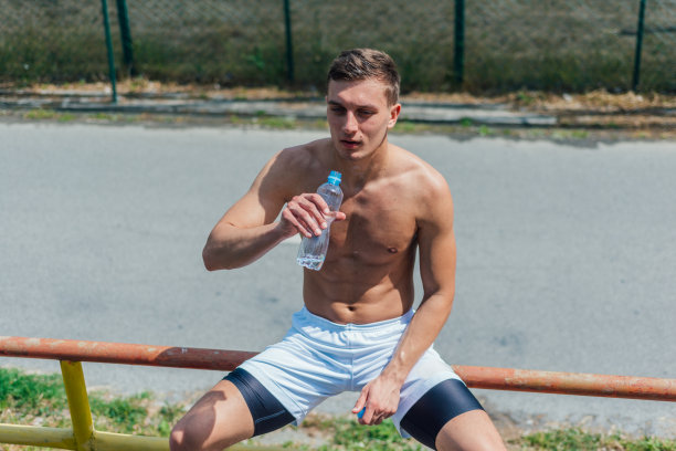 一个有魅力的男性运动员在锻炼完后坐在公园里，喝了一瓶水图片下载