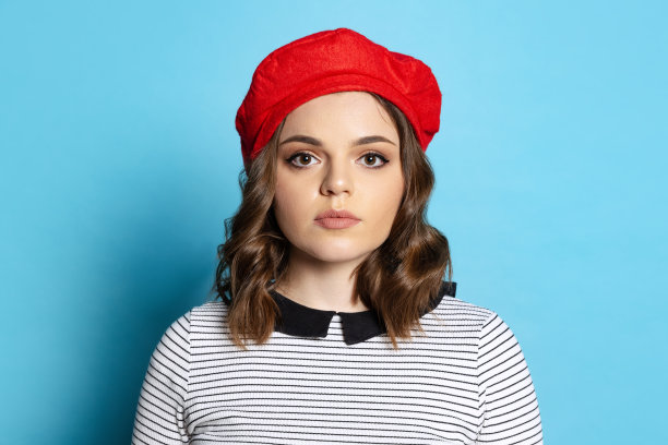年轻迷人的女孩的半身像与长卷发戴红色贝雷帽孤立在蓝色背景。情感、美、时尚的概念图片下载