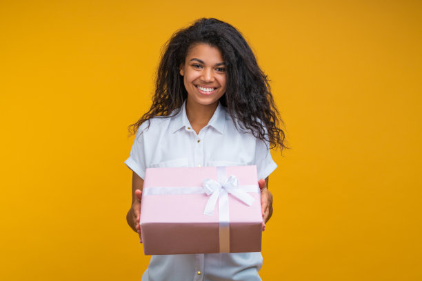 迷人的微笑时尚年轻女子的肖像摆造型与装饰礼品盒在手中，孤立在明亮的黄色背景图片下载