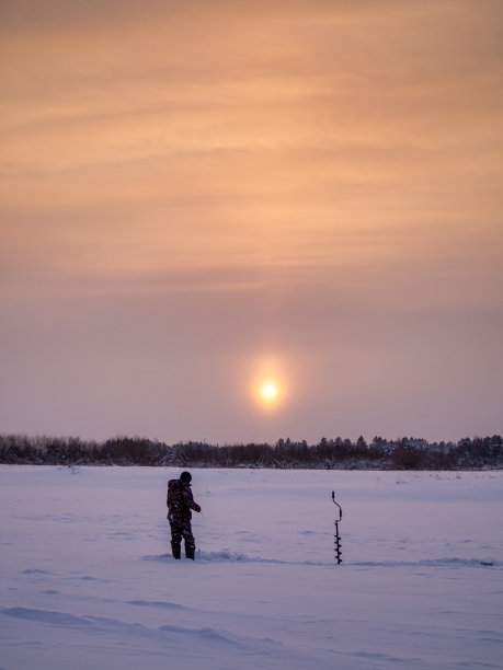 黎明时分，一个年轻人从冰上的一个洞里钓鱼出来。冬季钓鱼。图片下载