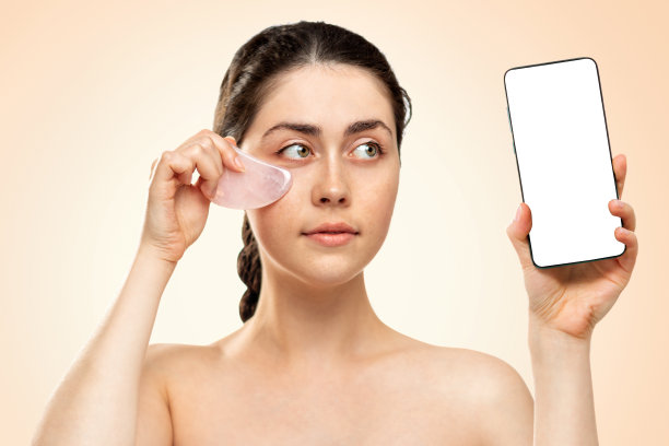 刮痧按摩。一名年轻女子一手拿着手机，一手拿着石榴石英石。模拟。米色背景。商业与美容的概念图片下载