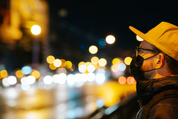 在散景灯前，一个戴着眼镜和面罩的男人的肖像。夜间拍摄的男性行人与城市街道散景背景图片下载