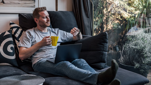 开朗微笑的青年男子舒舒服服地坐在家里的大沙发上，用笔记本电脑喝咖啡或喝茶，消磨空闲时间，看着手表图片下载