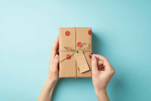 第一人称俯视图照片的情人节装饰女性手解开麻绳蝴蝶结在工艺纸礼品盒与圆点图案和标签在孤立的柔和的蓝色背景与copyspace图片下载