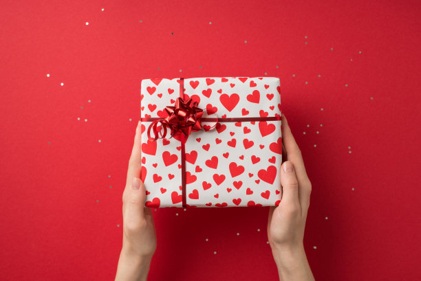第一人称俯视图照片的情人节装饰闪亮的亮片手拿礼品盒白色包装纸与心脏图案和红色星弓在孤立的红色背景与copyspace图片下载