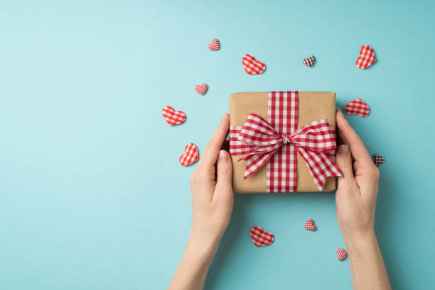 第一人称俯视图照片的情人节装饰手中的牛皮纸礼盒与格子丝带蝴蝶结包围装饰心在孤立的淡蓝色背景与空白空间图片下载