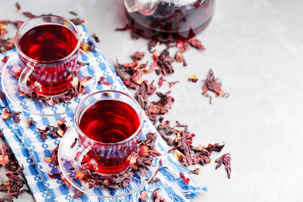 木槿茶和干燥的木槿花瓣在灰色的背景。两杯红色的芙蓉茶放在蓝色的餐巾上。前视图。本空间图片下载