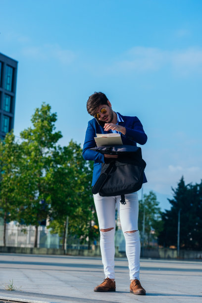 一个英俊的年轻商人站在户外，拿着文件，手机和公文包的全长照片。图片下载