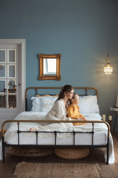 垂直生活方式照片的快乐年轻的母亲和女儿。充满柔情的妈妈和女婴坐在床上，拥抱在舒适的卧室与现代复古室内。快乐的母亲节图片下载