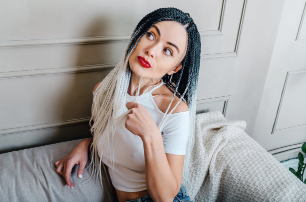 留着脏辫的性感女人。手靠近脸，神情沉思。白色t恤和蓝色牛仔裤。坐在灰色的沙发上。工作室的照片。图片下载