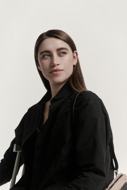 美丽的年轻女子的肖像与闪亮的黑色衬衫和人造毛皮米色包坐在白色背景的椅子美容化妆品保健概念图片下载