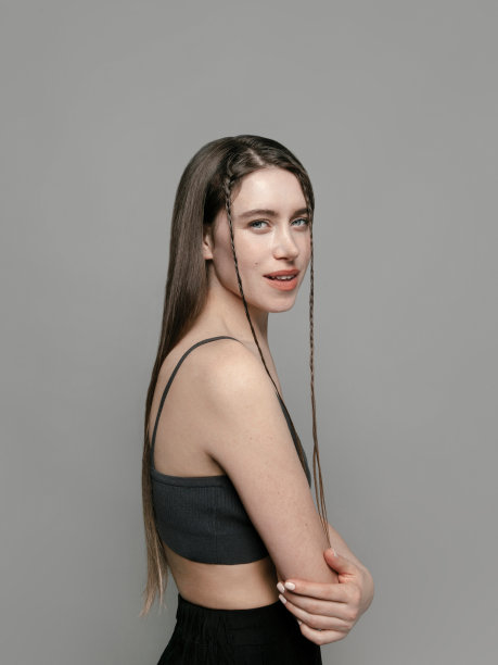 美丽的年轻女子的肖像与闪亮的化妆和辫子的头发穿着灰色的上衣和黑色的裤子在灰色的背景美容化妆品保健概念图片下载