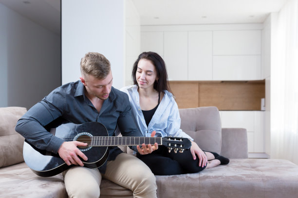 一对年轻夫妇，一男一女坐在家里的沙发上用笔记本电脑弹吉他，学习，上课，接吻，微笑图片下载