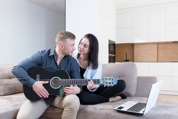 一对年轻夫妇，一男一女坐在家里的沙发上用笔记本电脑弹吉他，学习，上课，接吻，微笑图片下载