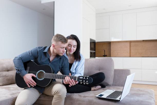 一对年轻夫妇，一男一女坐在家里的沙发上用笔记本电脑弹吉他，学习，上课，接吻，微笑图片下载