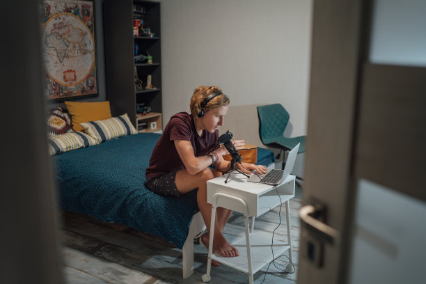 一个年轻的十几岁的男孩的肖像照片在耳机录音或聊天的朋友使用麦克风与流行滤镜和笔记本电脑坐在他的家庭房间的床上。图片下载