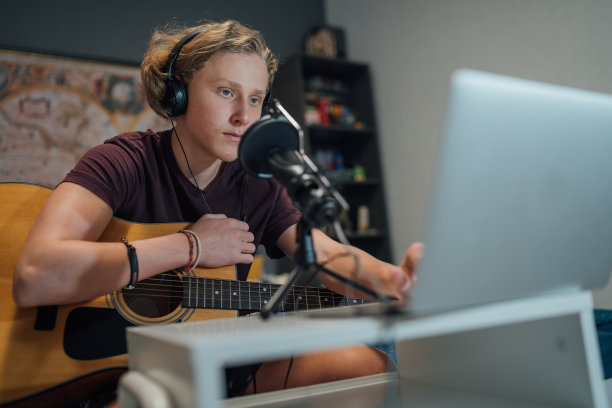 青少年的肖像在耳机里弹吉他浏览笔记本电脑。男孩用带滤镜的麦克风录音。现代音频录制技术概念图像。图片下载