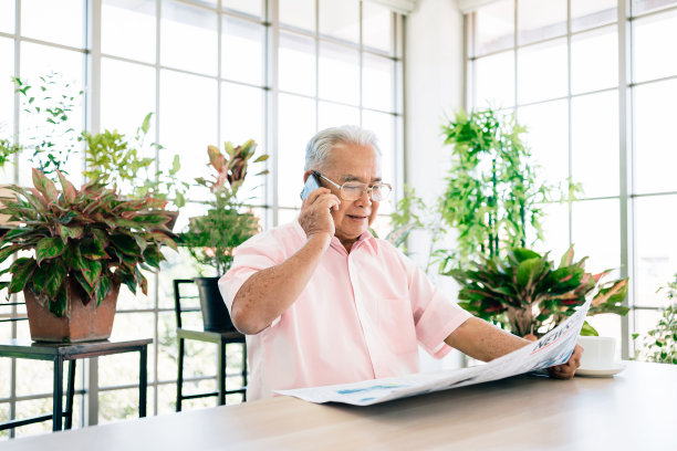 身穿粉红色休闲服的亚洲退休老人一边看报纸一边微笑着使用手机。坐在家里的室内花园里。退休爱好和生活方式。图片下载