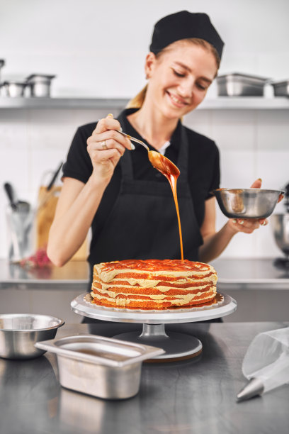 美丽可爱的女孩微笑着装饰蛋糕。穿着黑色制服的年轻糖果师在糖果店工作。图片下载