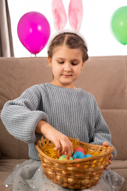 一个可爱的小女孩戴着兔耳朵头带，腿上放着柳条篮子，坐在房间的沙发上拿出一个复活节彩蛋图片下载